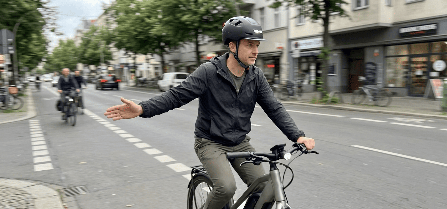 An adult commuter riding an eBike wearing an XNITO helmet on a city bike lane with one hand on the handlebar and the other signaling a turn